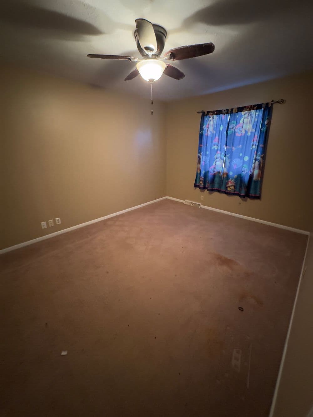 Empty room with beige walls, carpeted floor, ceiling fan, and a colorful curtain.