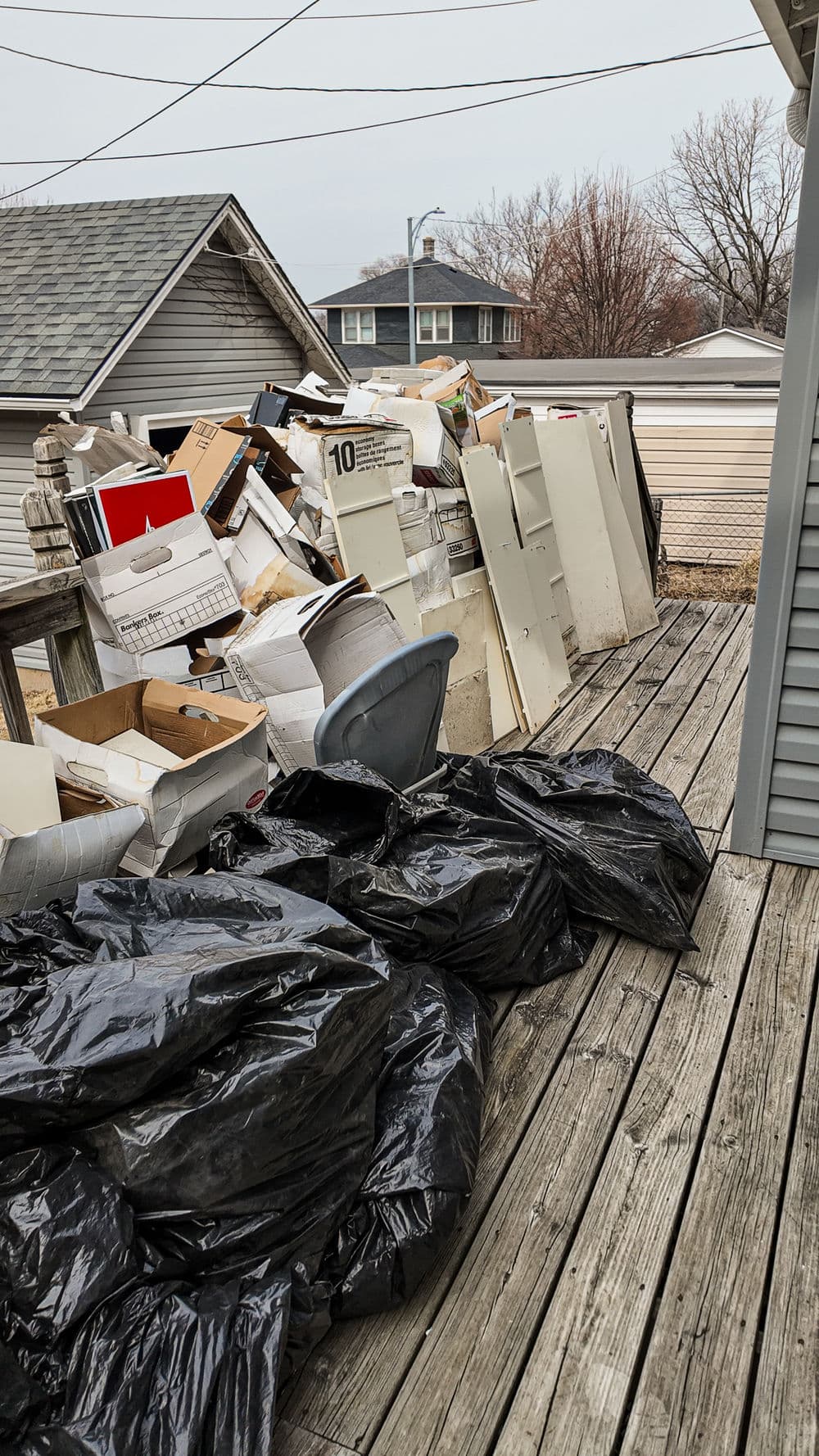 Debris and trash bags piled on a wooden deck with a gray house in the background.