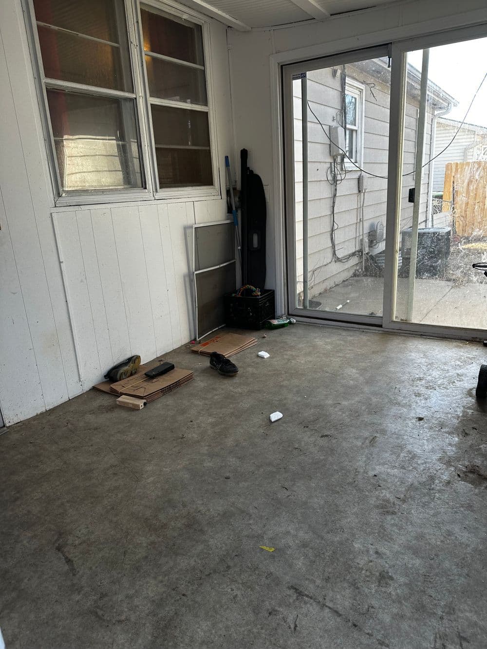 Empty porch with concrete floor, window view, and scattered items including shoes and a broom.
