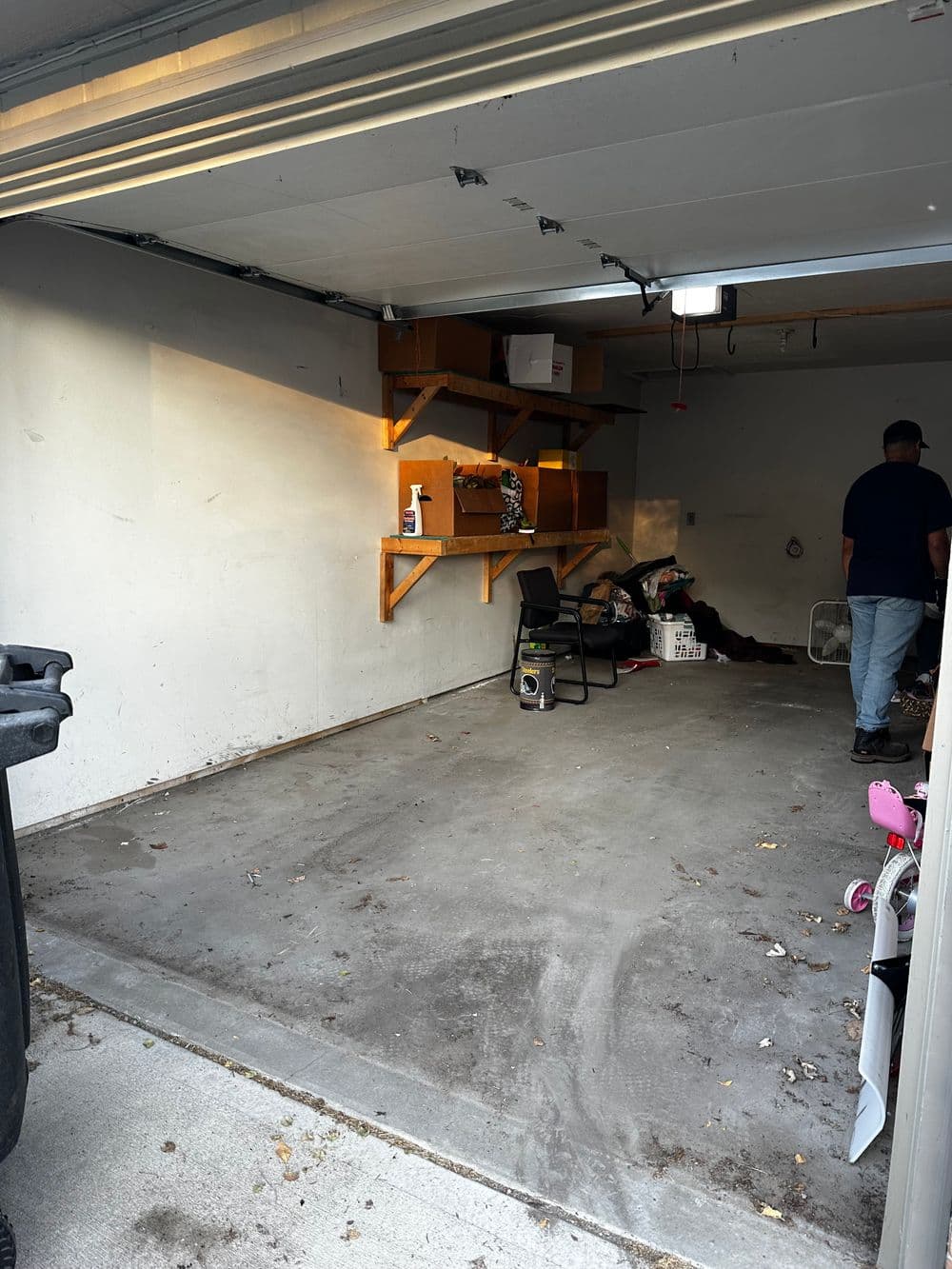 Empty garage with a chair, shelves, scattered items, and a trash can visible.