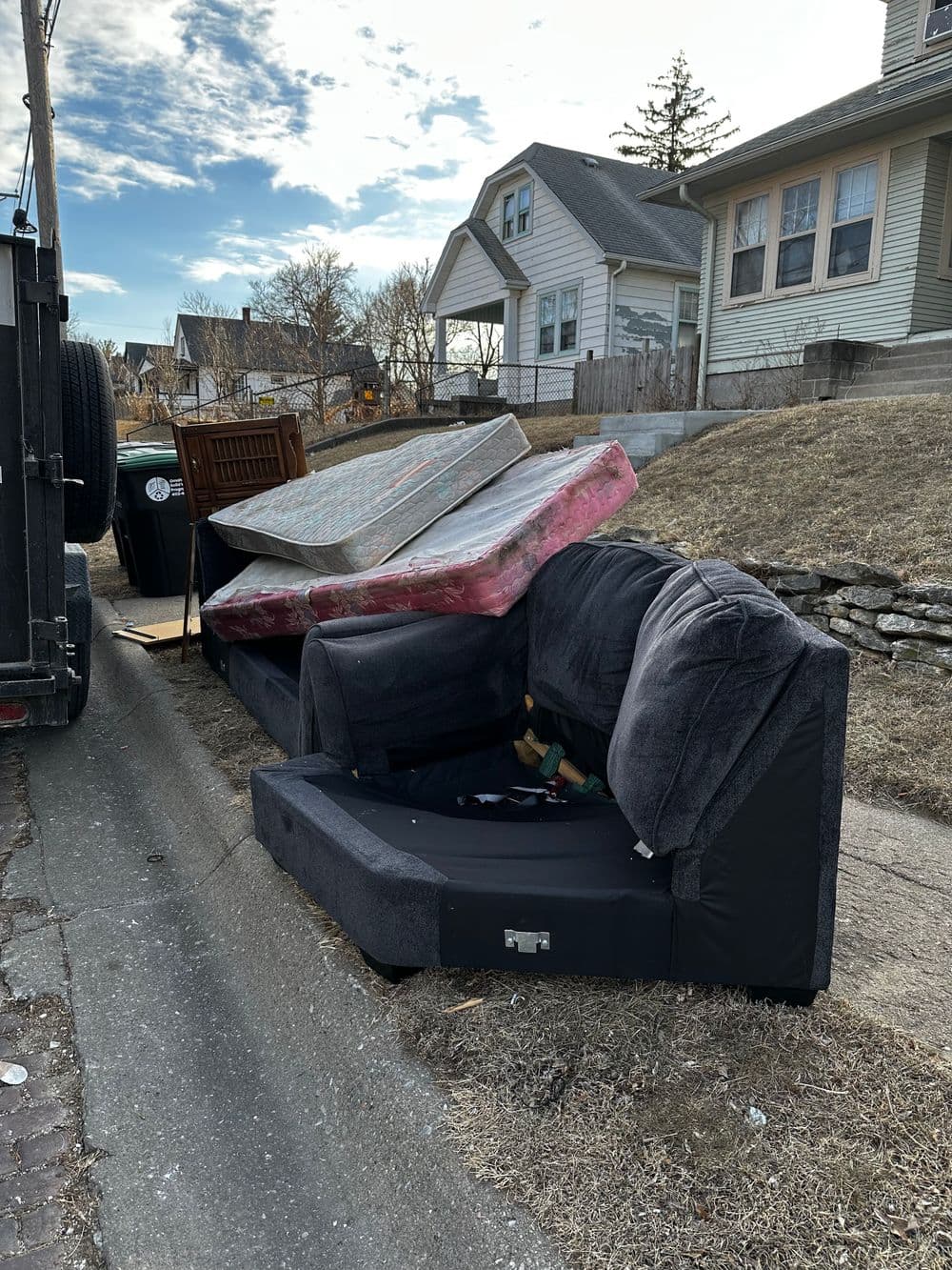 Abandoned black couch and mattresses by the curb on a residential street.