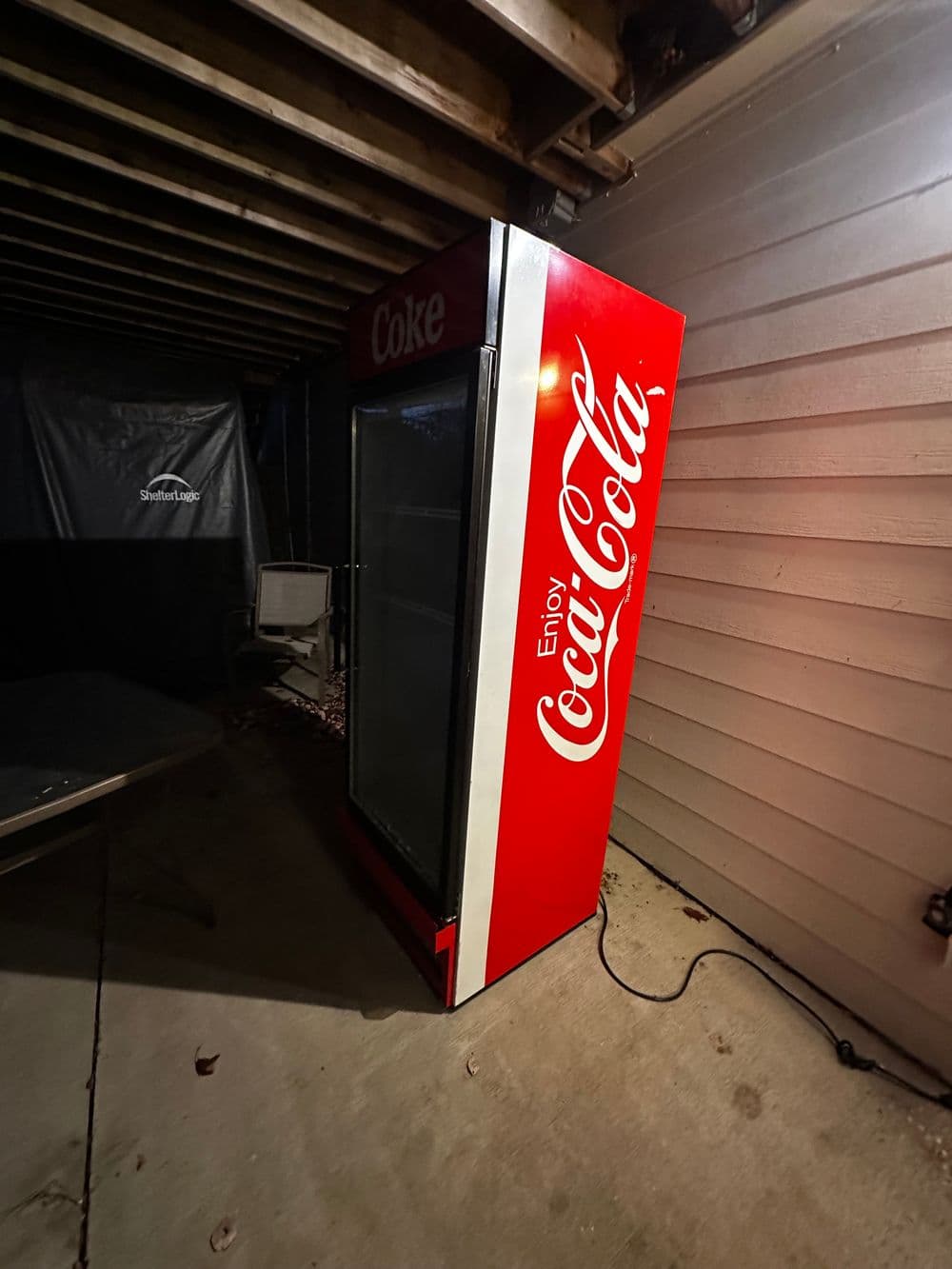 Coca-Cola vending machine in a dimly lit outdoor area, partially against a wall.