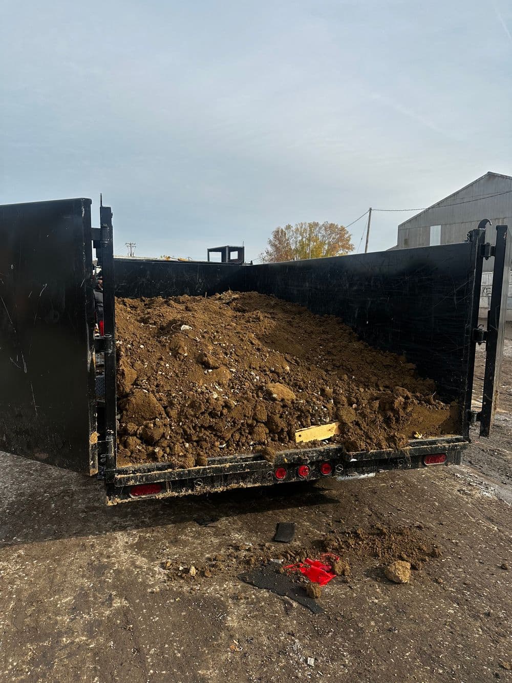 Dump trailer filled with dirt, parked on gravel surface with buildings in background.