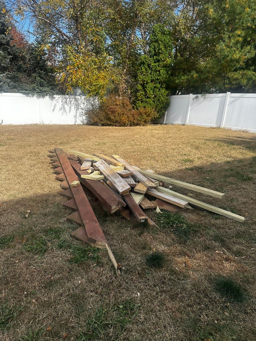 Piled wood planks and timber in a grassy yard with a white fence in the background.