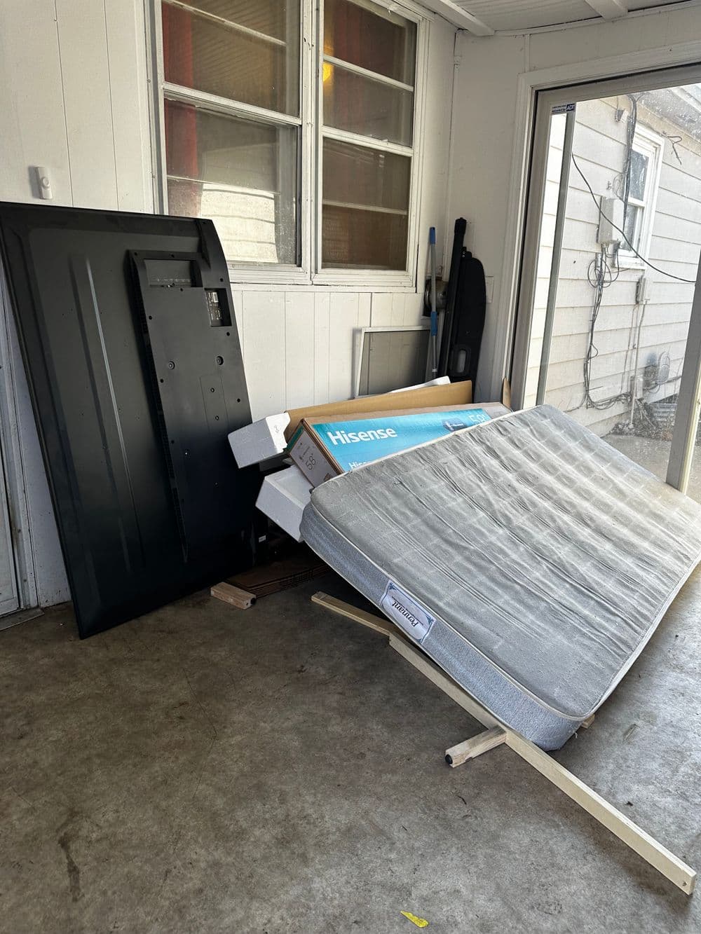 Mattress leaning against a wall with packaging materials and furniture in a cluttered space.