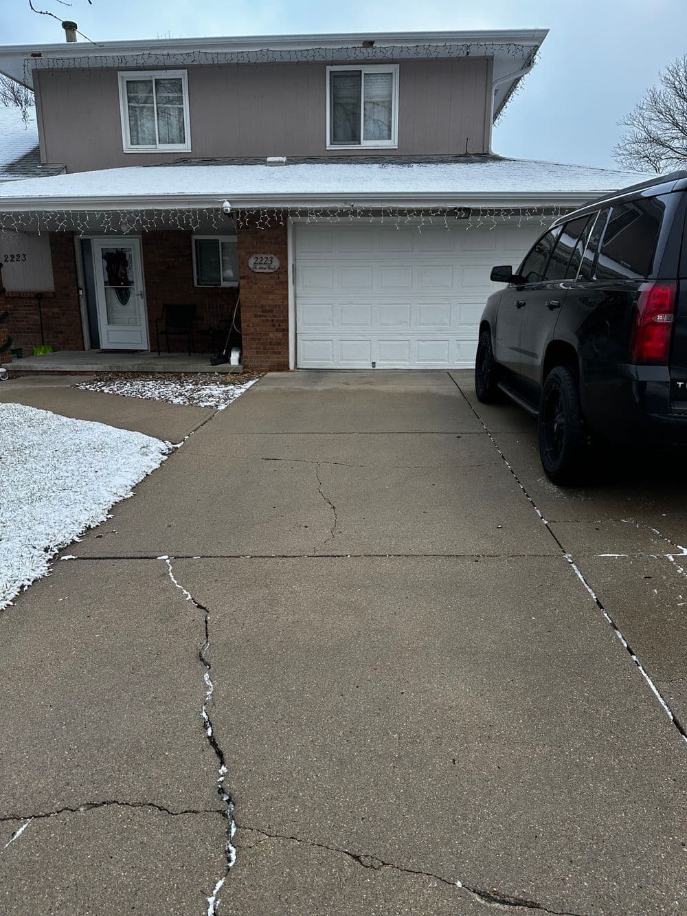 Snow-covered home with a cracked driveway and a black SUV parked nearby.