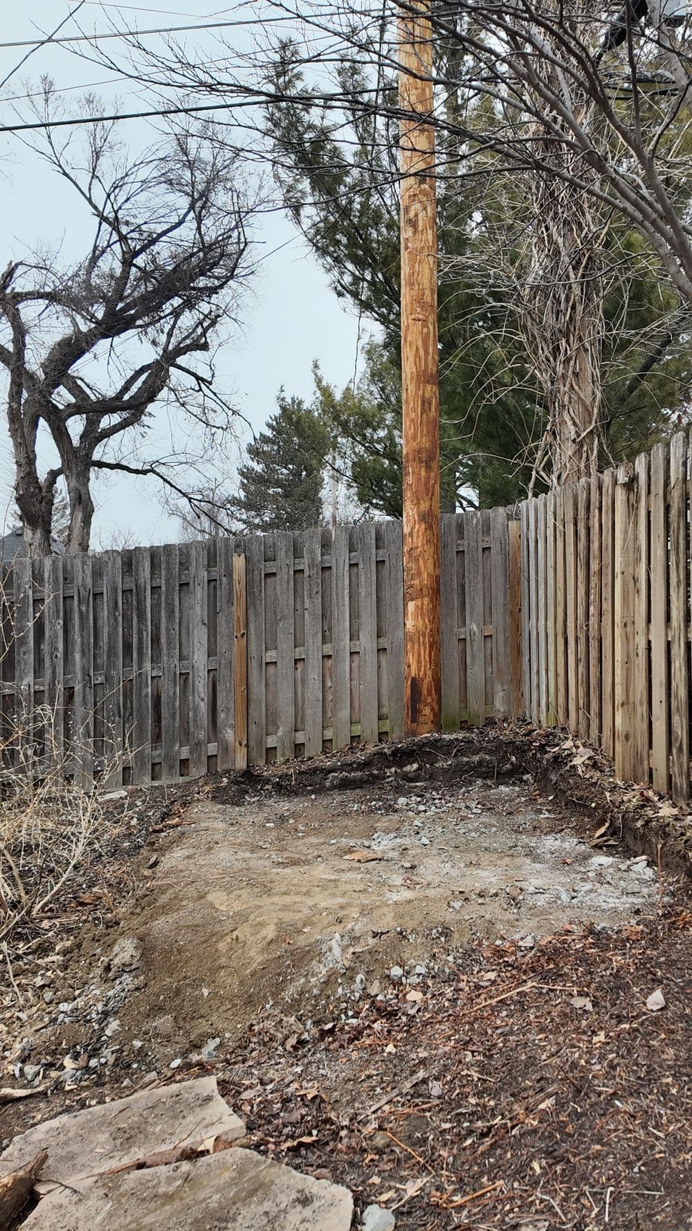 Vacant fenced area with exposed soil and a wooden utility pole surrounded by trees.