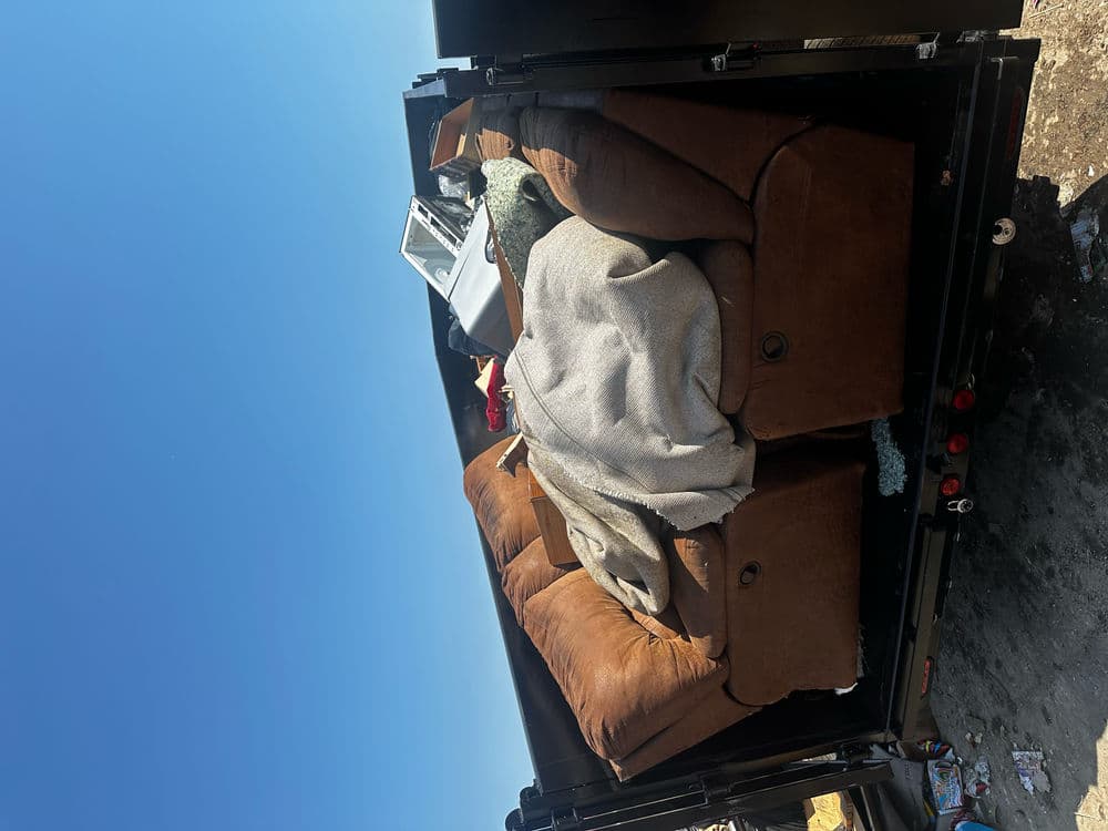 Brown couch with a blanket, discarded in a dumpster against a clear blue sky.