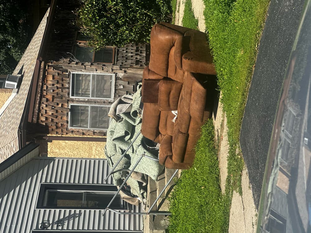 Brown leather couch stacked on grass next to a pile of rolled-up carpet.